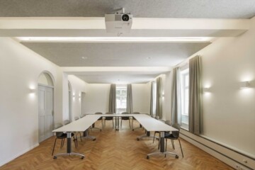 Heller Raum mit einem großen Tisch in U-Form. Links sind zwei große Türen. Geradezu und rechts erhellen mehrere deckenhohe Fenster den Raum.